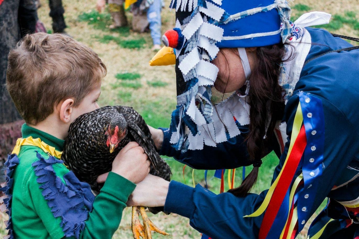 a woman in a colorful costume and mask hands a brown chicken to a young boy
