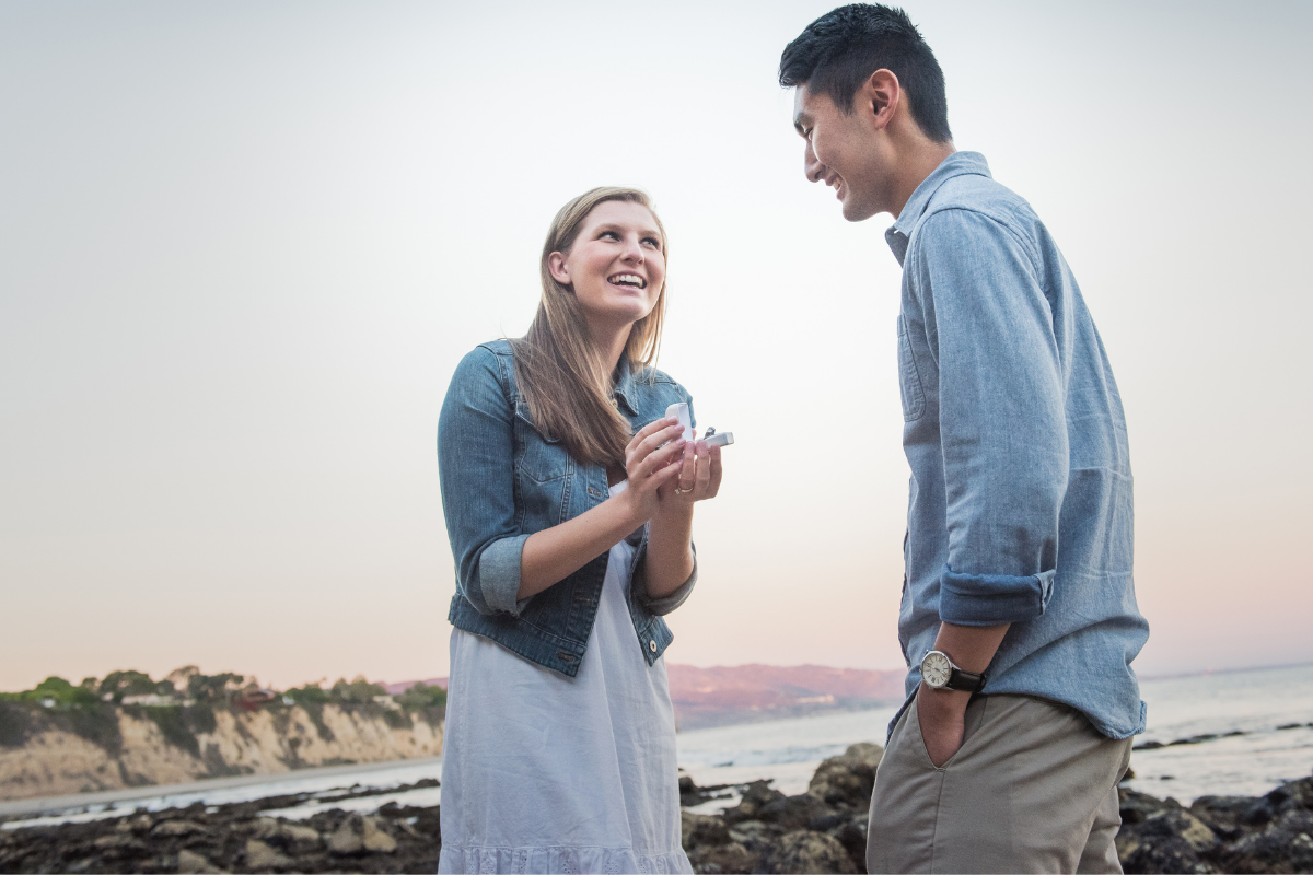 Caucasian young woman proposing with a diamond ring to his Japanese boyfriend at the beach during sunset.