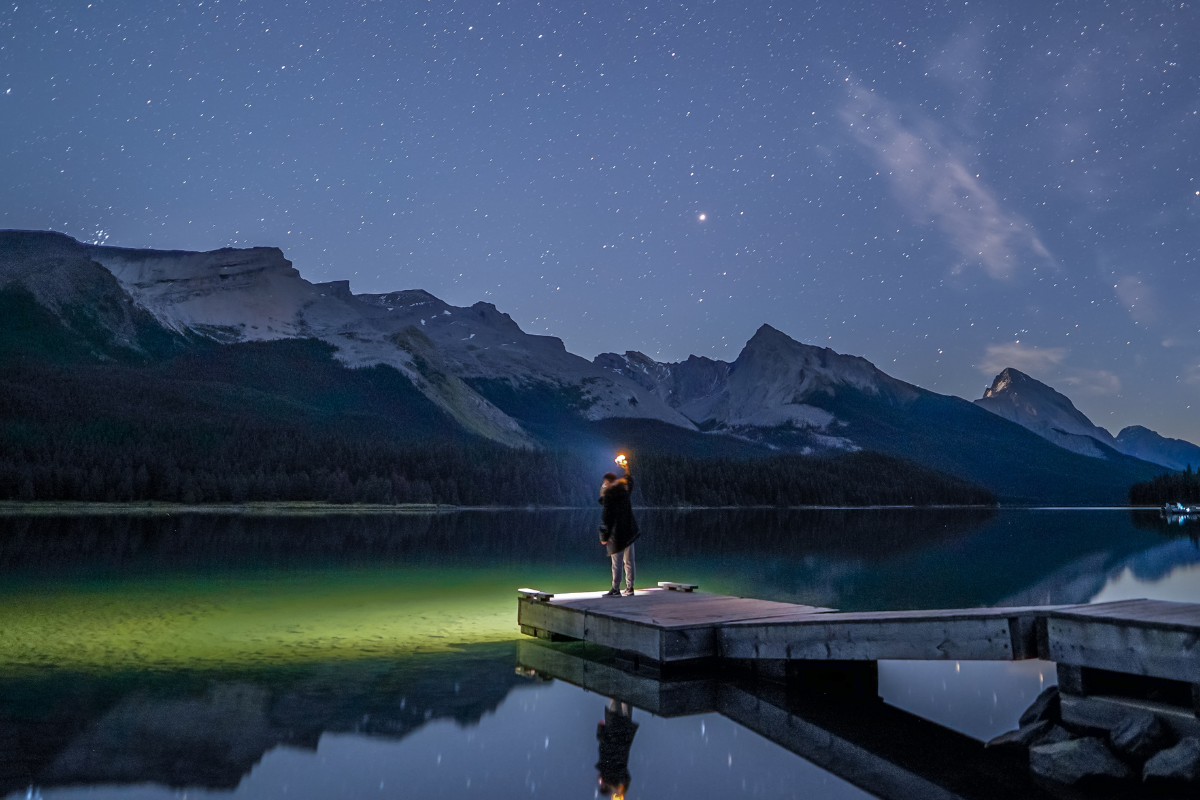 woman at edge of wooden pier shines flashlight on a lake with starry sky and mountains in background. winter