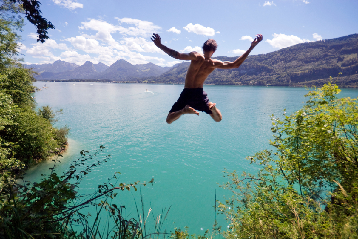 Young man cliff jumping into a blue Alpine lake below on a hot summer day.