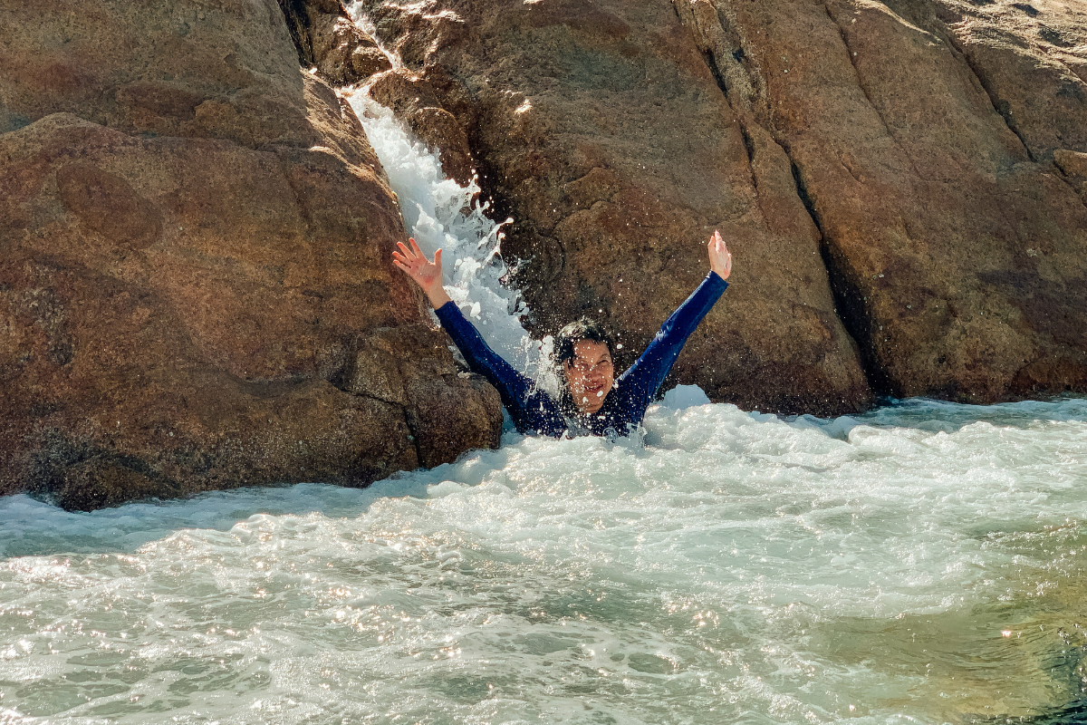 A shot of a mature asian woman coming down a natural rock slide into a pool of water. She is wearing a wetsuit and has her hands up in the air.